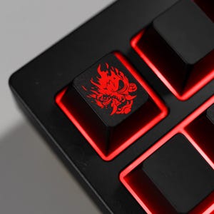 May include: A close-up of a black mechanical keyboard with red LED backlighting. The keycaps are black, with a red graphic of a stylized demon on one key. The keyboard is angled, showing the key layout and the red glow.