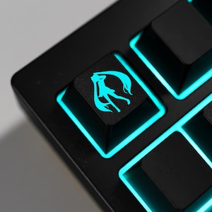 May include: Close-up of a black mechanical keyboard with teal backlighting. The keycaps are black, with a teal silhouette of a character on one key. The keyboard has a sleek, modern design, with the teal lighting adding a vibrant touch.