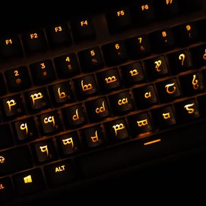May include: A black computer keyboard with backlit orange keys. The keycaps feature standard alphanumeric labels alongside Elvish Tengwar script characters. The layout includes function keys, a Windows key, and control keys, all illuminated with a warm glow.