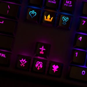 May include: A black computer keyboard featuring custom backlit keys with Kingdom Hearts symbols. The top row displays a blue heart with a crown, a yellow crown, and a blue heartless emblem. Below, pink keys show various heartless and nobody insignias.