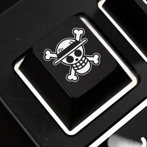 Pode incluir: Close-up de uma tecla de teclado preta com um design branco de caveira e ossos cruzados, com um chapéu de palha. A tecla faz parte de um teclado preto com acabamento branco. O design é um símbolo popular de anime e manga.