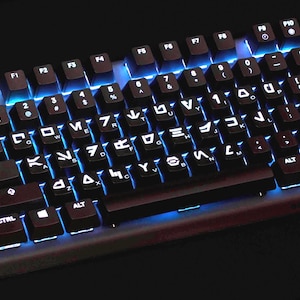 Puede incluir: Un teclado mecánico negro para computadora con teclas personalizadas que presentan caracteres Aurebesh. Las teclas están retroiluminadas con una luz azul vibrante que ilumina los espacios entre los interruptores, resaltando las letras únicas.
