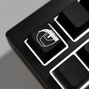 May include: Close-up of a black keyboard with white key outlines. One key features a white outline of a helmet. The keyboard keys are arranged in a grid pattern. The image is well-lit, highlighting the details of the keyboard.