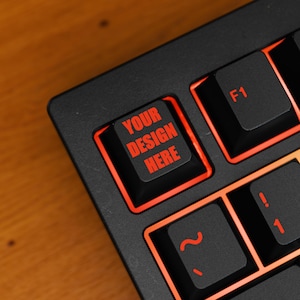 Peut inclure: Gros plan sur un clavier noir avec des touches éclairées en rouge. Les touches affichent des lettres rouges, dont "YOUR DESIGN HERE" et "F1". Le clavier est incliné, montrant une partie des touches et le cadre.