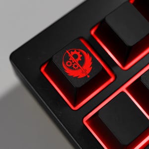 Op de afbeelding: Close-up van een zwart mechanisch toetsenbord met rode LED-achtergrondverlichting. De keycap heeft een rode afbeelding van een tandwiel, zwaard en vleugels. Het toetsenbord is ontworpen voor gaming en heeft een strakke, moderne uitstraling.