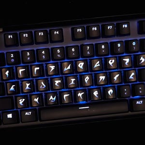 May include: A black computer keyboard featuring backlit keys with custom white symbols instead of standard letters. The keys are illuminated by a bright blue light emanating from beneath the keycaps, highlighting the unique characters.
