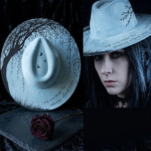 May include: A light blue fedora hat with a black weeping willow tree design and the text "She can make the weeping willows smile." A dark red rose rests on a slate surface.