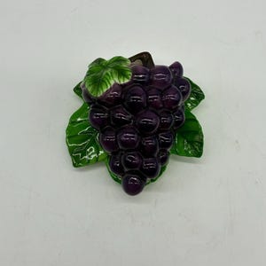 May include: A ceramic grape cluster with deep purple grapes and bright green leaves. The leaves have white vein details. The grapes are clustered together, and the overall design is a decorative piece.