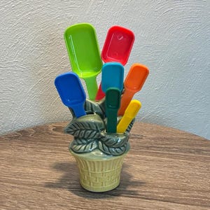 May include: A ceramic flower pot holds a collection of colorful scoops. The scoops are in various bright colors, including green, red, blue, orange, and yellow. The pot is a light yellow with a basket weave design and leaf accents.