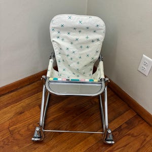 May include: Vintage baby rocker with a white seat featuring a teal star pattern. The chair has a chrome frame, a tray, and a footrest. The rocker is designed with a retro aesthetic, perfect for a nursery or vintage decor.