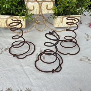 May include: Three decorative, rusty brown metal springs in a spiral shape. The springs are on a white surface with floral embroidery. In the background, a gold-colored metal plant stand holds three cream-colored planters filled with green foliage.
