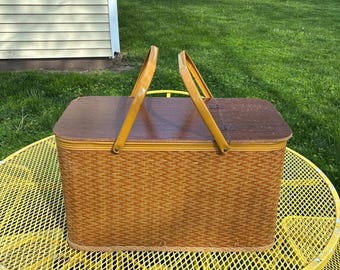 May include: A vintage picnic basket with a woven brown body, a dark wooden lid, and a yellow handle. The basket is rectangular and sits on a yellow metal table, perfect for outdoor dining.