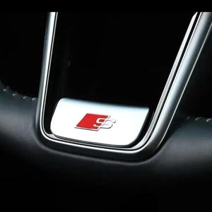 Peut inclure: Un volant en cuir noir doté d'un insert en argent métallique à la base. L'insert affiche un emblème S-line rouge et argent avec la lettre S dans une police stylisée. Le volant présente des coutures visibles le long du bord incurvé.