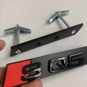 Puede incluir: Emblema SQ5 negro y cromado con un acento rojo, junto con el hardware de montaje. El emblema presenta las letras "SQ5" en una fuente audaz y estilizada. El hardware de montaje incluye soportes metálicos negros y tornillos plateados.