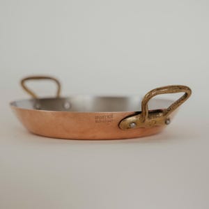 Puede incluir: Una sartén Mauviel 1830 de color cobre con dos asas de latón. La sartén tiene un interior plateado y el texto "MAUVIEL 1830 Made in France" está impreso en el lateral.