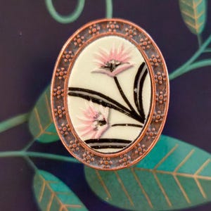 May include: An oval-shaped brooch with a floral design. The brooch features a white background with pink flowers and black stems, framed by a copper-colored border. The brooch is set against a dark blue background with green leaves.