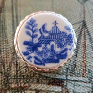 May include: A round, white and blue porcelain button with a silver-colored metal rim. The button features a blue and white Willow pattern design. The button is set against a patterned background with a nautical theme.