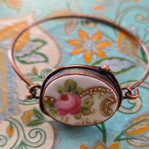 May include: A copper-toned bangle bracelet with an oval, porcelain centerpiece. The centerpiece features a pink rose and green leaves on a cream background, accented with gold detailing. The bracelet is set against a floral patterned background.