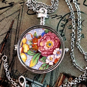 May include: A silver chain necklace with a round pendant featuring a floral design in shades of pink, yellow, blue, and green. The pendant is made from a vintage china plate.