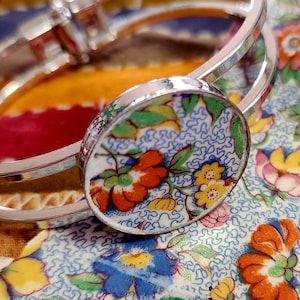 May include: A silver-toned bangle bracelet with a circular, floral-patterned charm. The charm features orange, yellow, and blue flowers on a light blue background. The bracelet rests on a floral fabric with a mix of colorful blooms.