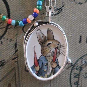 May include: Silver pendant necklace featuring a Peter Rabbit illustration. The oval pendant has a white background with a rabbit wearing a blue jacket and holding two carrots. A beaded necklace with colorful beads is also visible.