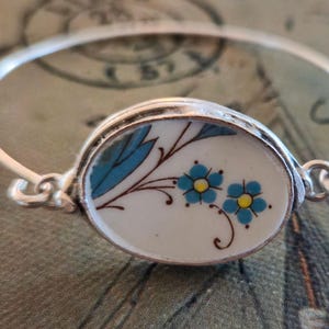 May include: A silver-toned metal bangle bracelet featuring a central oval-shaped ceramic or porcelain inlay. The white inlay displays a painted design of two blue flowers with yellow centers, green leaves, and thin brown stems with delicate swirls.