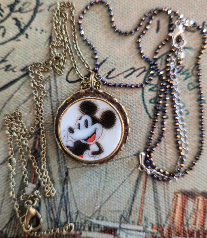 Op de afbeelding: Een ronde hangerketting met een afbeelding van Mickey Mouse op een witte achtergrond, gevat in een getextureerd bronskleurig metalen frame. De hanger is bevestigd aan een dunne, donker metalen bolletjesketting met een karabijnsluiting.