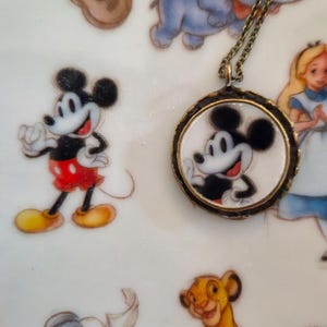 Op de afbeelding: Een ronde hangerketting met een zwart-witte illustratie van Mickey Mouse op een witte achtergrond, gevat in een donker metalen frame en bevestigd aan een dunne ketting, rustend op een bord versierd met diverse illustraties van stripfiguren.