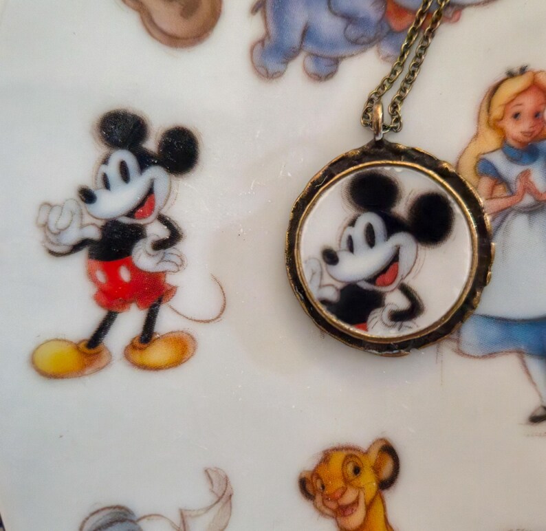 Op de afbeelding: Een ronde hangerketting met een zwart-witte illustratie van Mickey Mouse op een witte achtergrond, gevat in een donker metalen frame en bevestigd aan een dunne ketting, rustend op een bord versierd met diverse illustraties van stripfiguren.