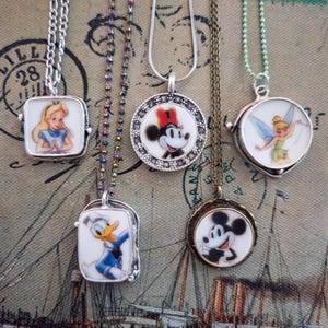 Op de afbeelding: Vijf kettingen met ronde en rechthoekige metalen hangers met kleurrijke illustraties van stripfiguren, waaronder Alice, Donald Duck, Minnie Mouse, Mickey Mouse en Tinker Bell, elk hangend aan verschillende soorten metalen kettingen.