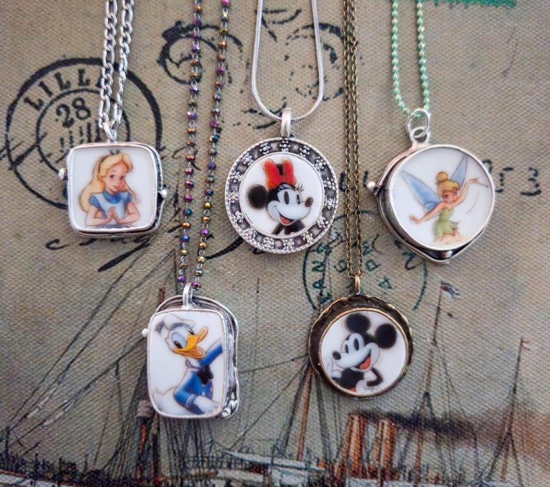 Op de afbeelding: Vijf kettingen met ronde en rechthoekige metalen hangers met kleurrijke illustraties van stripfiguren, waaronder Alice, Donald Duck, Minnie Mouse, Mickey Mouse en Tinker Bell, elk hangend aan verschillende soorten metalen kettingen.