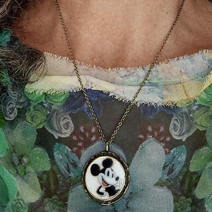 Op de afbeelding: Een ronde hangerketting met een zwart-witte illustratie van Mickey Mouse op een witte achtergrond, gevat in een donker metalen frame. De hanger hangt aan een dunne, donker bronskleurige metalen ketting die om de nek rust.