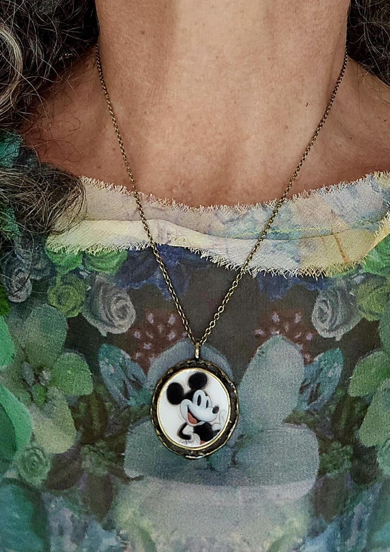 Op de afbeelding: Een ronde hangerketting met een zwart-witte illustratie van Mickey Mouse op een witte achtergrond, gevat in een donker metalen frame. De hanger hangt aan een dunne, donker bronskleurige metalen ketting die om de nek rust.