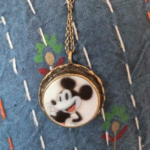 Op de afbeelding: Een ronde hangerketting met een zwart-witte illustratie van Mickey Mouse op een witte achtergrond, gevat in een getextureerd, donker metalen frame, hangend aan een dunne, bijpassende schakelketting van donker metaal.