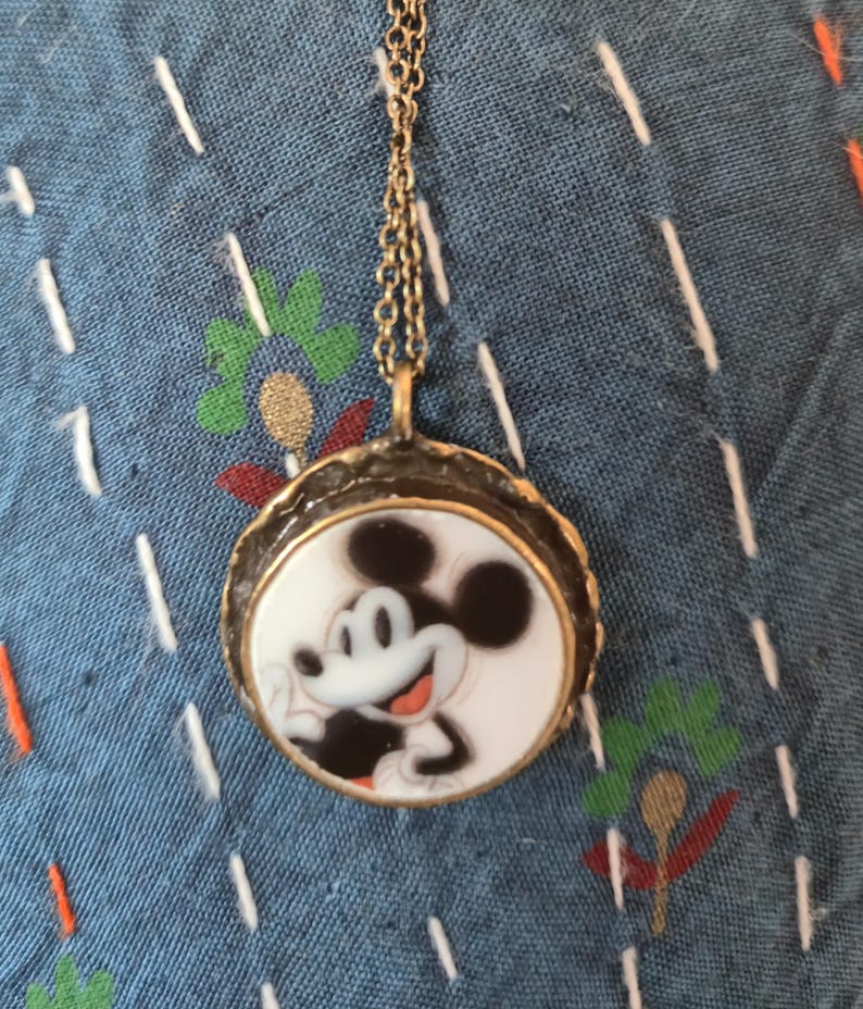 Op de afbeelding: Een ronde hangerketting met een zwart-witte illustratie van Mickey Mouse op een witte achtergrond, gevat in een getextureerd, donker metalen frame, hangend aan een dunne, bijpassende schakelketting van donker metaal.