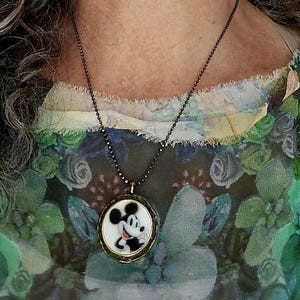 Op de afbeelding: Een ronde hangerketting met een zwart-witte illustratie van Mickey Mouse op een witte achtergrond, gevat in een donker metalen frame. De hanger hangt aan een dunne, donkergekleurde kralenketting die op de borst rust.