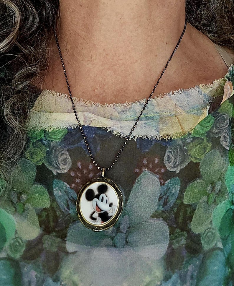 Op de afbeelding: Een ronde hangerketting met een zwart-witte illustratie van Mickey Mouse op een witte achtergrond, gevat in een donker metalen frame. De hanger hangt aan een dunne, donkergekleurde kralenketting die op de borst rust.