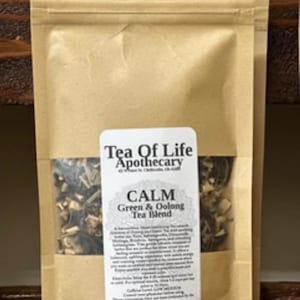 May include: A brown paper bag of "Tea Of Life Apothecary" Calm Green & Oolong Tea Blend. The label includes the text "Calm" and "Green & Oolong Tea Blend". The bag contains dried tea leaves and herbs.