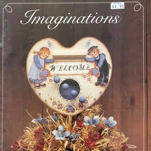 May include: A heart-shaped wooden sign with the word "WELCOME" painted on it. Two children are painted on the sign, one holding a blue bucket and the other holding a red bucket. The sign is decorated with flowers and blue balls. The sign is sitting on a basket of flowers.