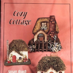 May include: Three decorative cottage figurines with moss-covered roofs and charming details. The cottages feature red doors, small windows, and floral accents. The image includes the text "Cozy Cottage" and the names "Susan Roach" and "Emily Dinsdale".