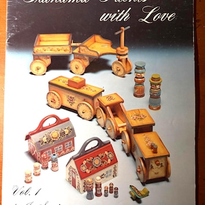 May include: A book cover titled Grandma Paints with Love Vol. 1 by Jo Sonja featuring illustrations of hand-painted wooden toys including a wagon, a train, two small house-shaped containers, and several small wooden figures with floral designs.