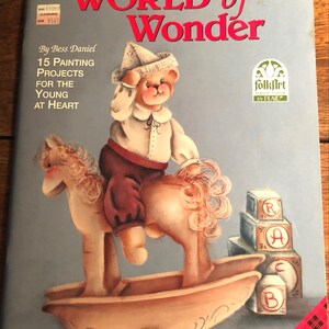 May include: A book cover titled "WORLD of Wonder" featuring a painted illustration of a teddy bear riding a rocking horse. The cover also includes text that reads "15 PAINTING PROJECTS FOR THE YOUNG AT HEART" and the Plaid and FolkArt logos.