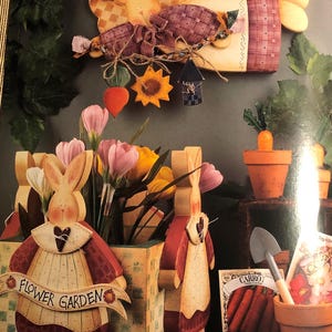 May include: A wooden flower garden planter featuring two rabbit figures wearing aprons, a wall-mounted carrot-shaped sign with a plaid pattern, a small terracotta pot with a carrot, a metal trowel, and a packet of carrot seeds.