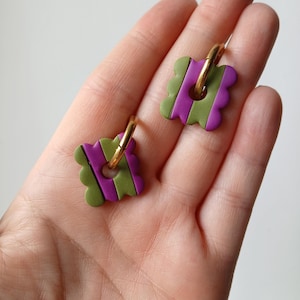 Peut inclure: Une paire de boucles d'oreilles créoles avec des pendentifs carrés aux bords festonnés. Les pendentifs présentent des rayures verticales alternées vert olive et violet vif, avec une découpe circulaire centrale où passe un anneau en métal doré.