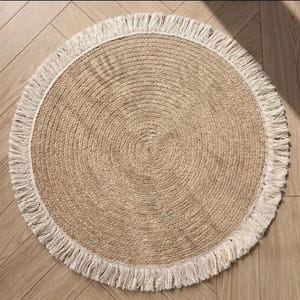 May include: A round jute rug with a natural tan color and a white fringe border. The rug has a woven texture and is suitable for indoor use. The rug's diameter is approximately 36 inches.