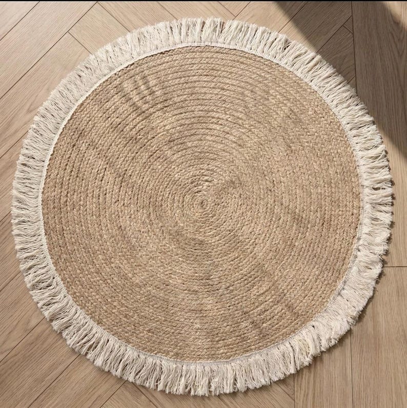 May include: A round jute rug with a natural tan color and a white fringe border. The rug has a woven texture and is suitable for indoor use. The rug's diameter is approximately 36 inches.