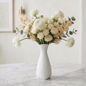 May include: A white, textured vase filled with a bouquet of white and peach-colored flowers. The arrangement includes roses and other blooms, with green foliage. The vase sits on a marble-patterned surface, creating a fresh, elegant display.