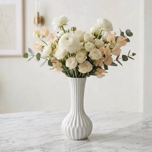Peut inclure: Un vase en céramique blanc, au design cannelé, contient un bouquet de fleurs blanches et pêche. L'arrangement comprend des roses, des renoncules et d'autres fleurs, avec des accents de feuillage vert. Le vase est posé sur une surface à motifs de marbre.