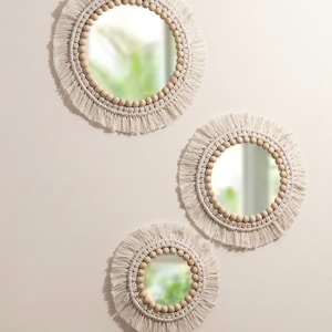 May include: Three round macrame mirrors of varying sizes. Each mirror has a circular glass center, surrounded by a ring of wooden beads and a fringe of off-white macrame. The mirrors are mounted on a light-colored wall.