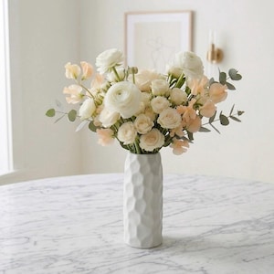 Könnte beinhalten: Eine weiße Keramikvase mit geometrischem Muster enthält einen Strauß weißer und pfirsichfarbener Blumen. Das Arrangement umfasst Rosen, Ranunkeln und andere Blüten, mit grünen Blättern. Die Vase steht auf einem Tisch mit Marmorplatte.